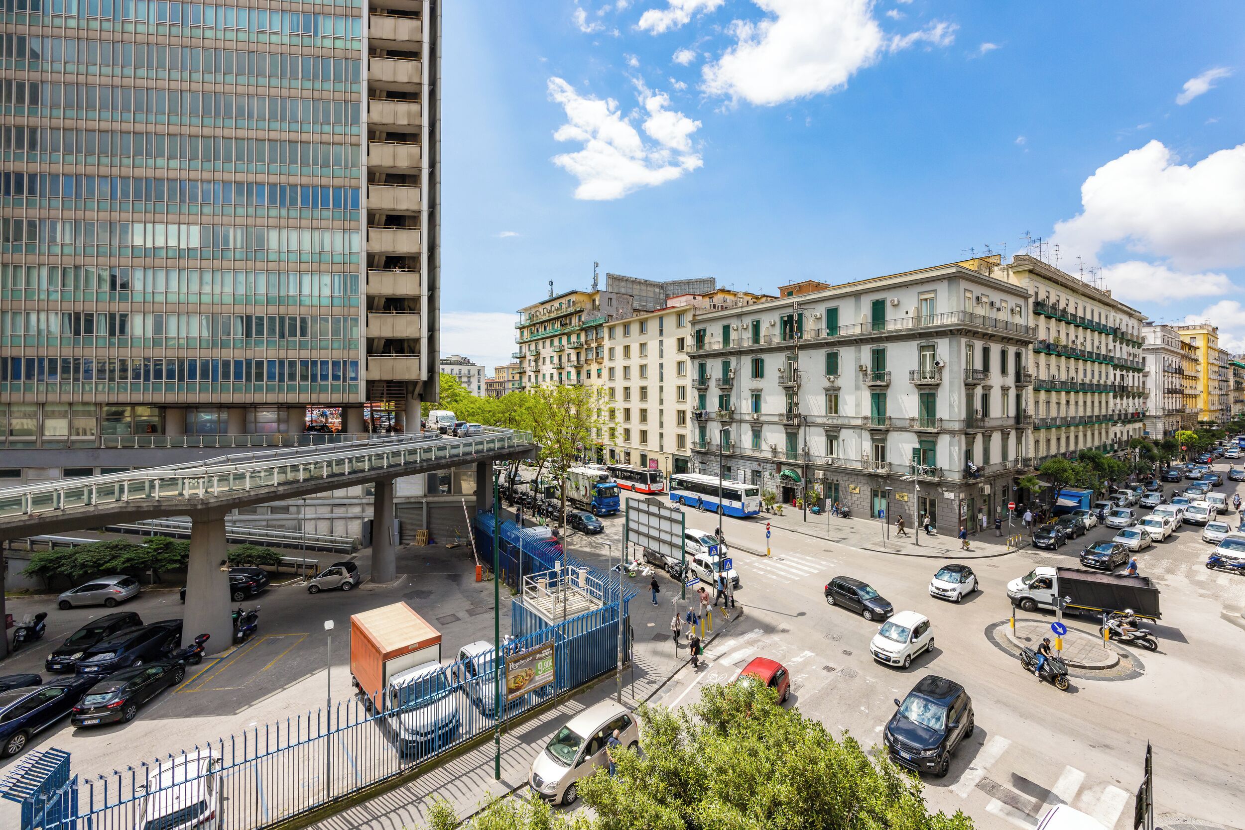 Een drukke stadstraat met een voetgangersbrug, geparkeerde auto's en gebouwen met klassieke architectuur.