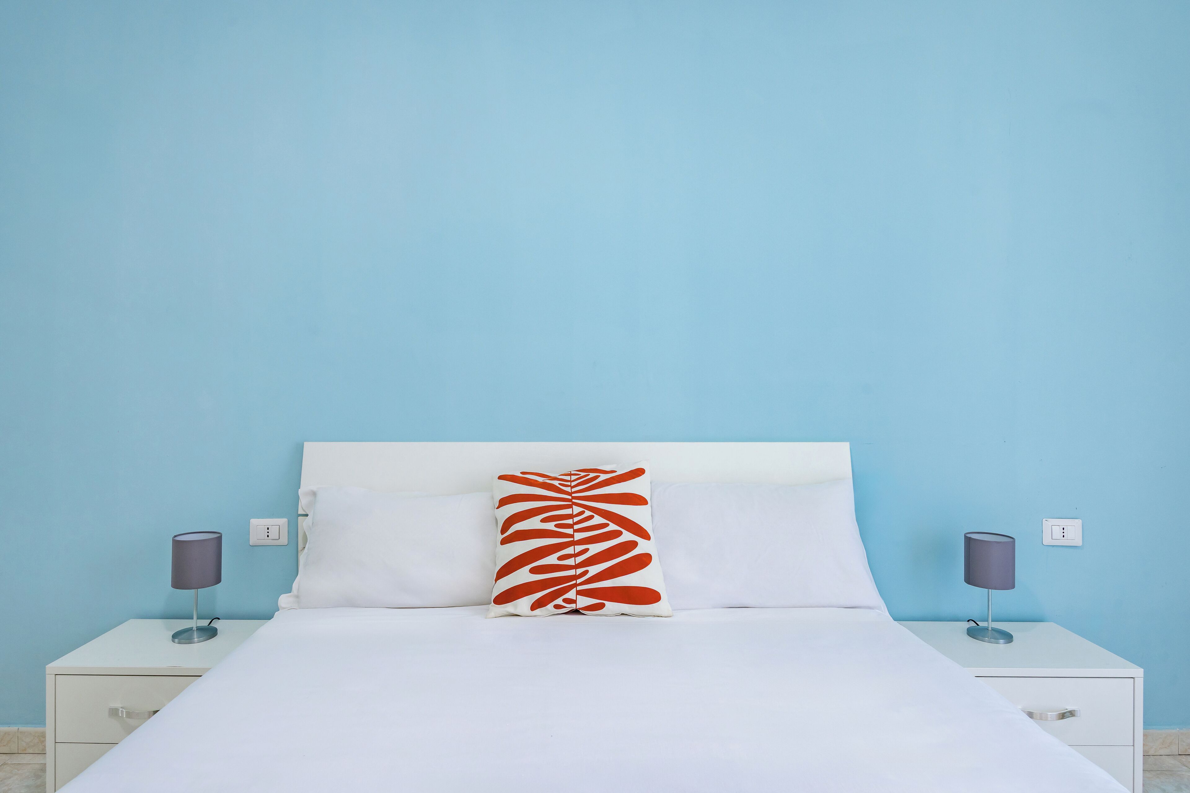 A bed with white bedding, a white headboard, and a red and white pillow. There are two bedside tables with lamps and a power outlet on the wall.