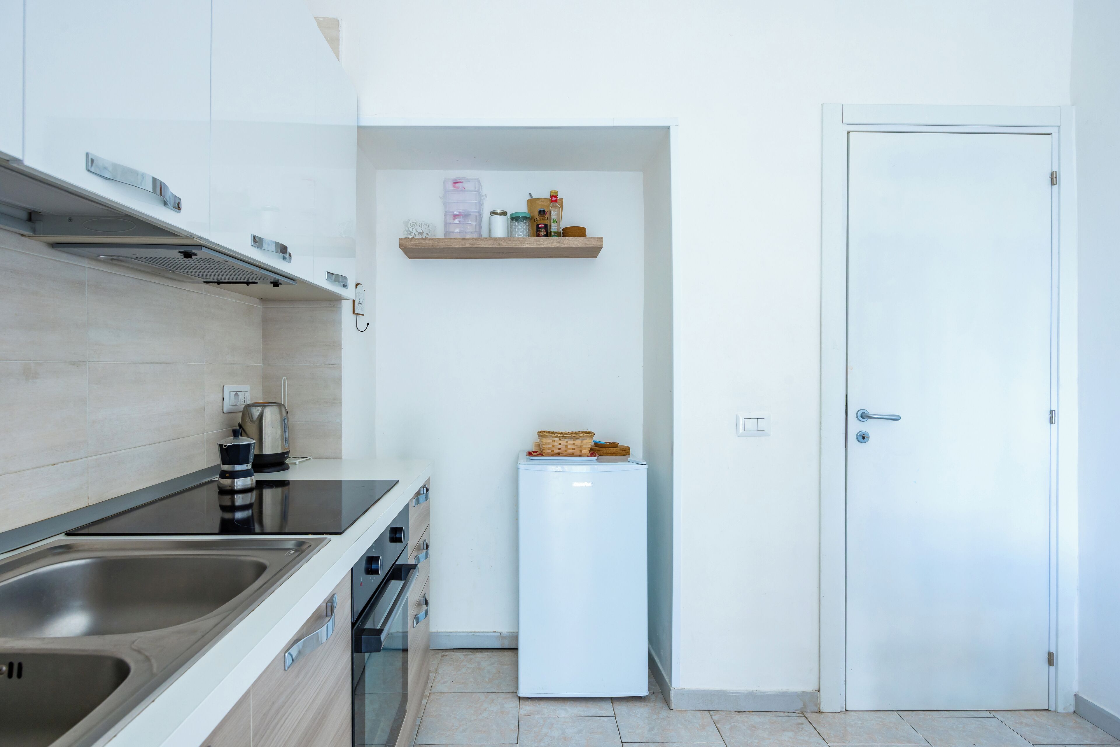 Une cuisine compacte équipée d’un évier à deux bacs, d’une cuisinière et d’un réfrigérateur. Il y a une petite étagère au-dessus du réfrigérateur et une porte sur la droite.