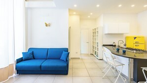 Una sala de estar moderna con un sofá azul, sillas blancas, una zona de cocina con un armario amarillo y una decoración de mariposa en la pared.