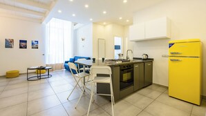Una cocina moderna con un refrigerador amarillo, una zona de comedor con mesa y sillas, y un sofá azul en una habitación luminosa con ventanas amplias.