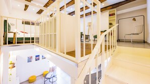 Un loft espacioso con vigas de madera a la vista, una escalera blanca y un entrepiso con asientos y obras de arte.