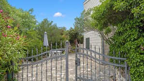 A gated courtyard with a stone floor, surrounded by lush greenery and a building with a light-colored facade. - Amazing home in Rogoznica with WiFi (Rogoznica)