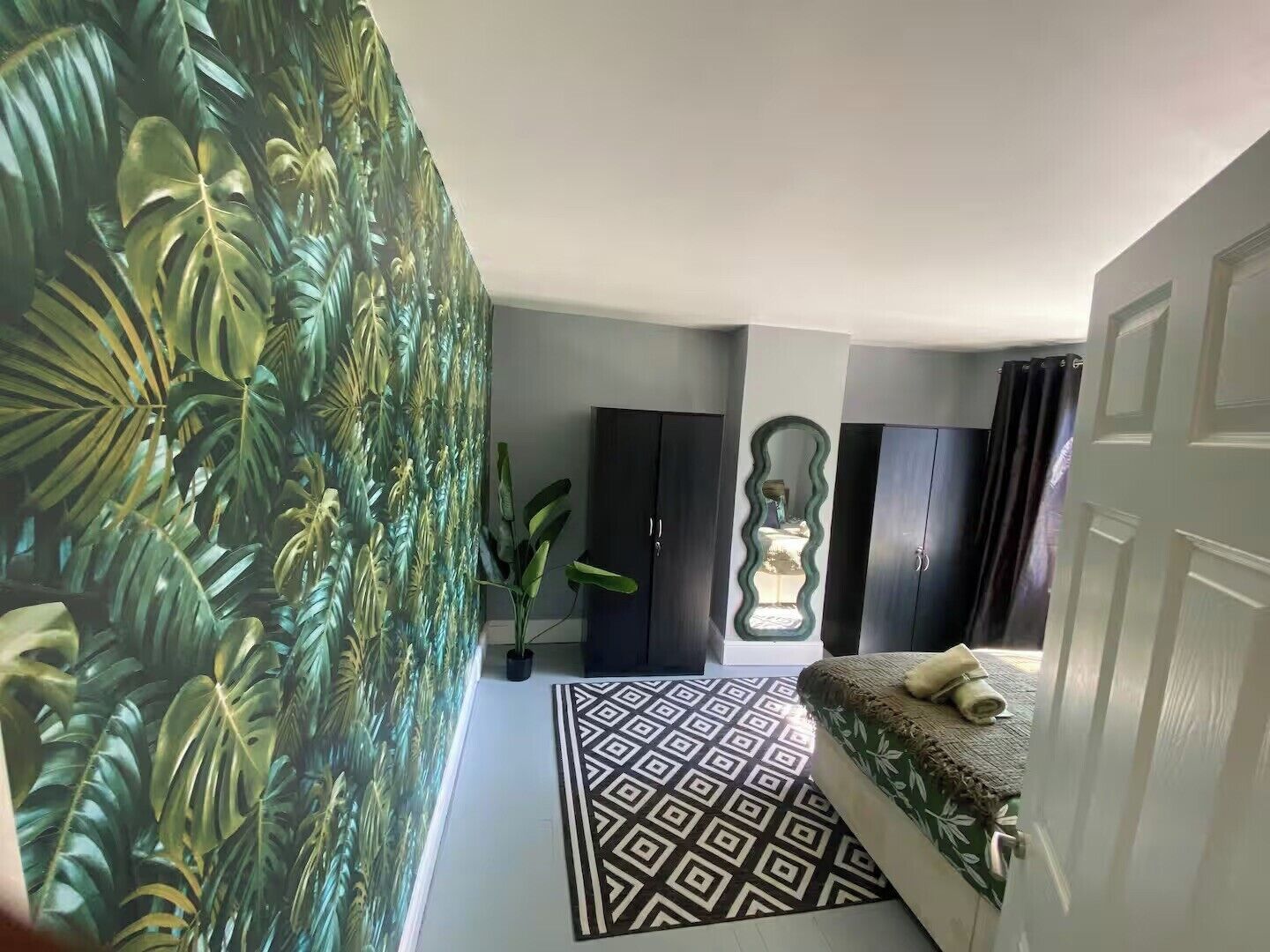 A bedroom with a patterned rug, a bed with a green and white floral bedspread, a potted plant, and a wall with a tropical leaf wallpaper.