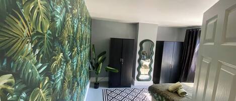 A bedroom with a patterned rug, a bed with a green and white floral bedspread, a potted plant, and a wall with a tropical leaf wallpaper.