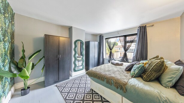 A bedroom with a bed, a wardrobe, a mirror, a potted plant, and a patterned rug. - 4 Bd House - Sleeps 8 With Garden & Parking (Gillingham)