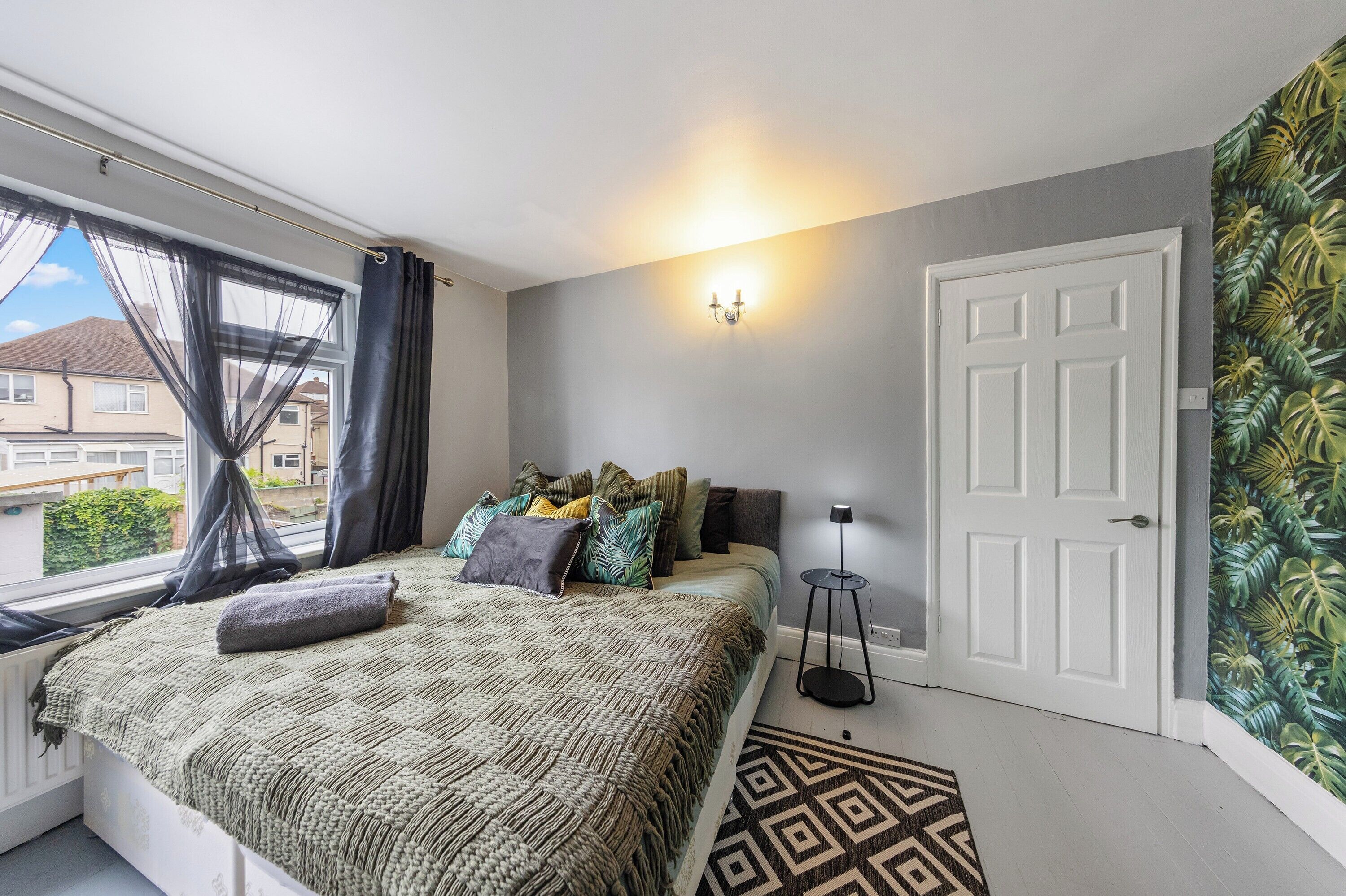 A bedroom with a bed, a window with curtains, a wall-mounted light, a door, and a patterned rug.