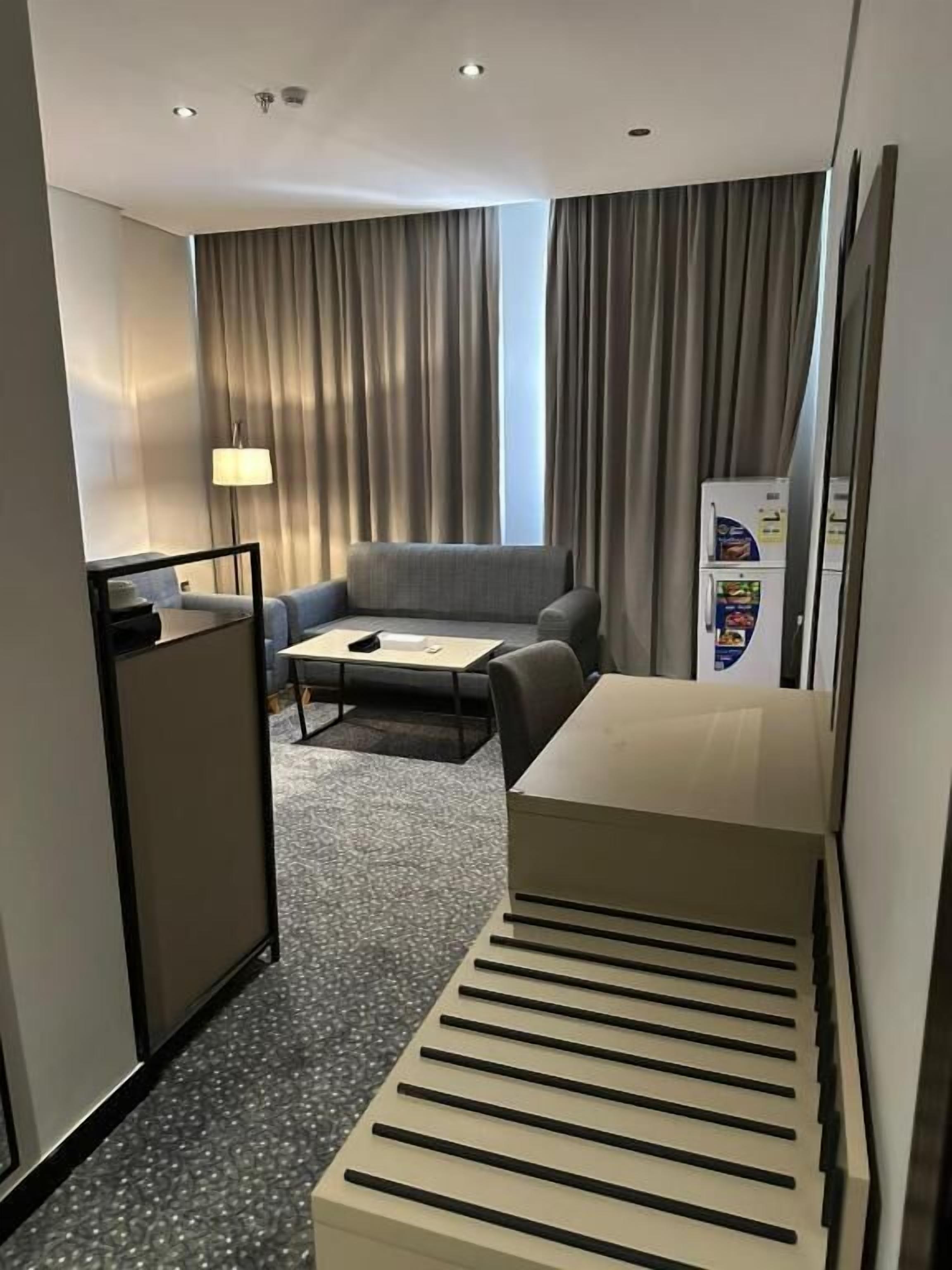 A compact hotel room with a sofa, a small table, a desk, a refrigerator, and a patterned carpet.