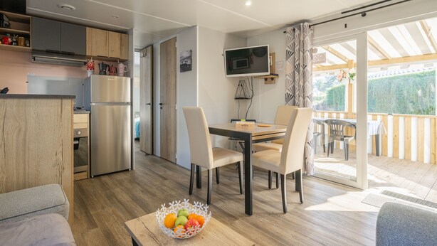 A modern kitchen with stainless steel appliances, a dining area with a table and chairs, and a balcony with a wooden deck. - Bora-Bora Mobile Home 289 with Shared Pool, Private Terrace and Garden (Les mathes)