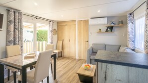 A modern living room with a dining area, wooden flooring, and a built-in kitchen counter. - Bora-Bora Mobile Home 289 with Shared Pool, Private Terrace and Garden (Les mathes)