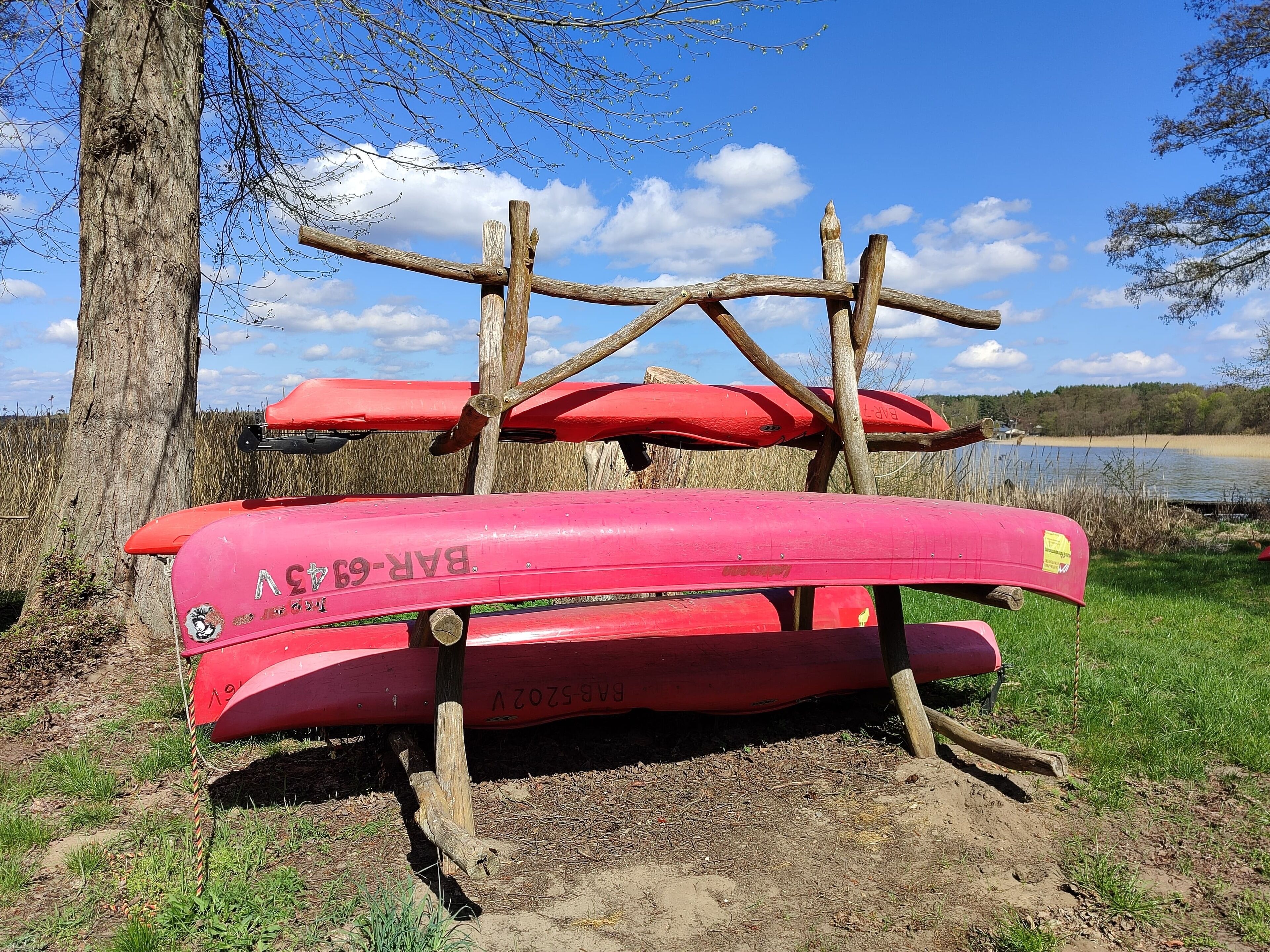 Una canoa roja con texto visible y una estructura de madera que la sostiene, frente a un fondo de árboles y una masa de agua.