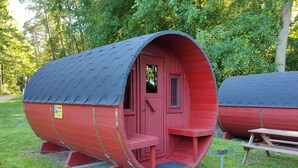 A red cylindrical structure with a door and windows, a bench, and a picnic table labeled "Cupid". - TinyHouse "Cupid" by the lake, Mecklenburg Lake District (Fürstenberg/Havel)