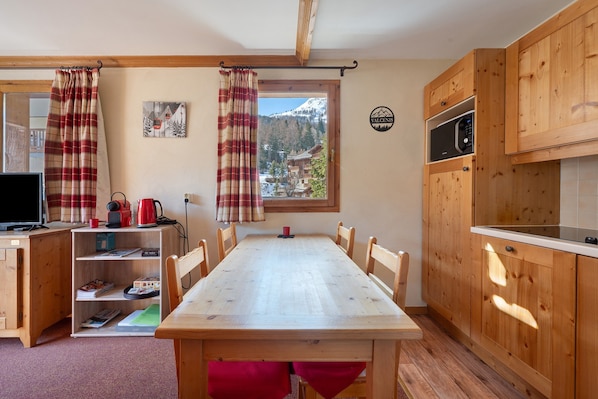 A wooden dining table with chairs, a kitchen with wooden cabinets, a microwave, and a view of a snowy mountain through a window.
