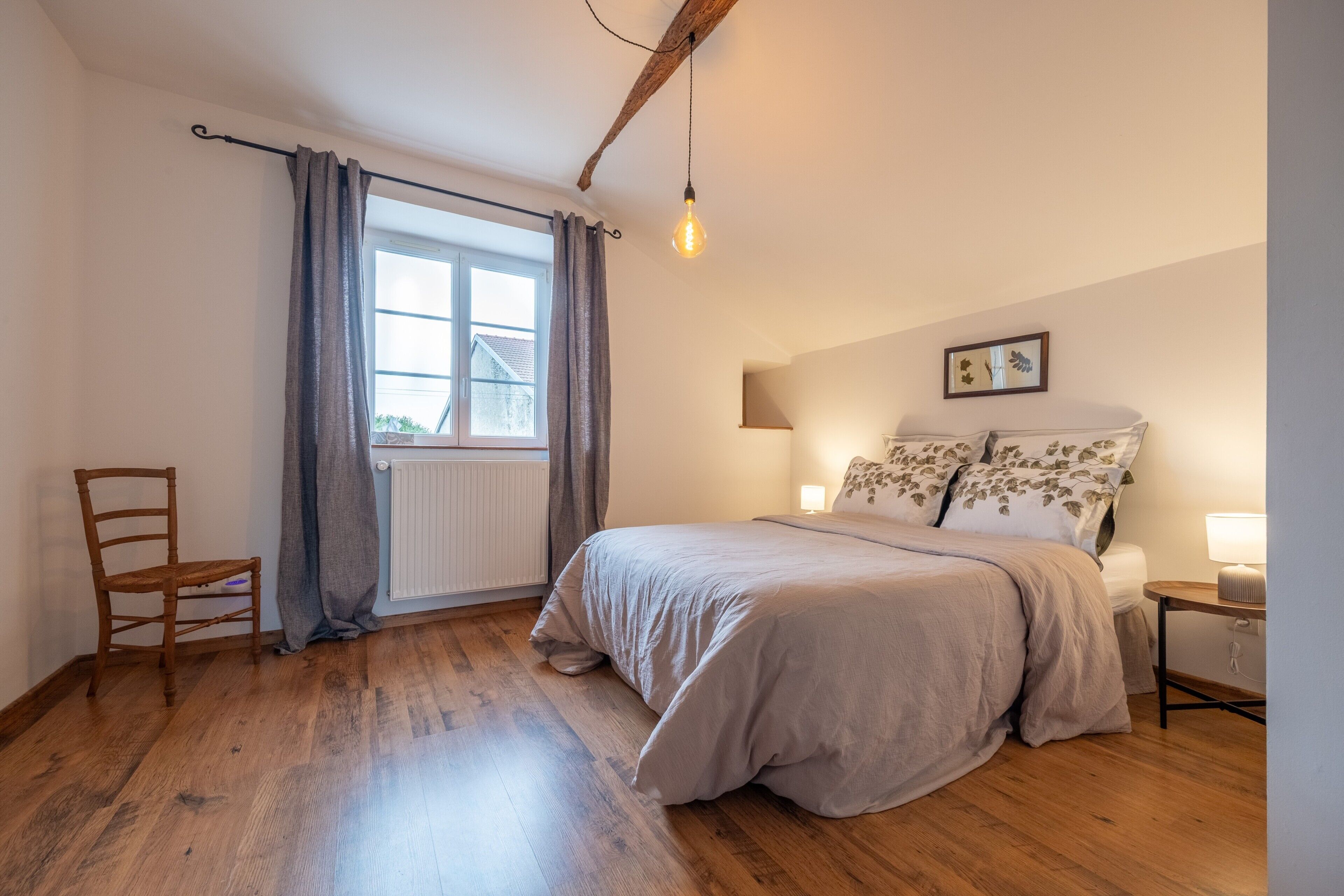 A bedroom with a wooden floor, a bed with a floral pillow set, a chair, a window with curtains, and a hanging light bulb.