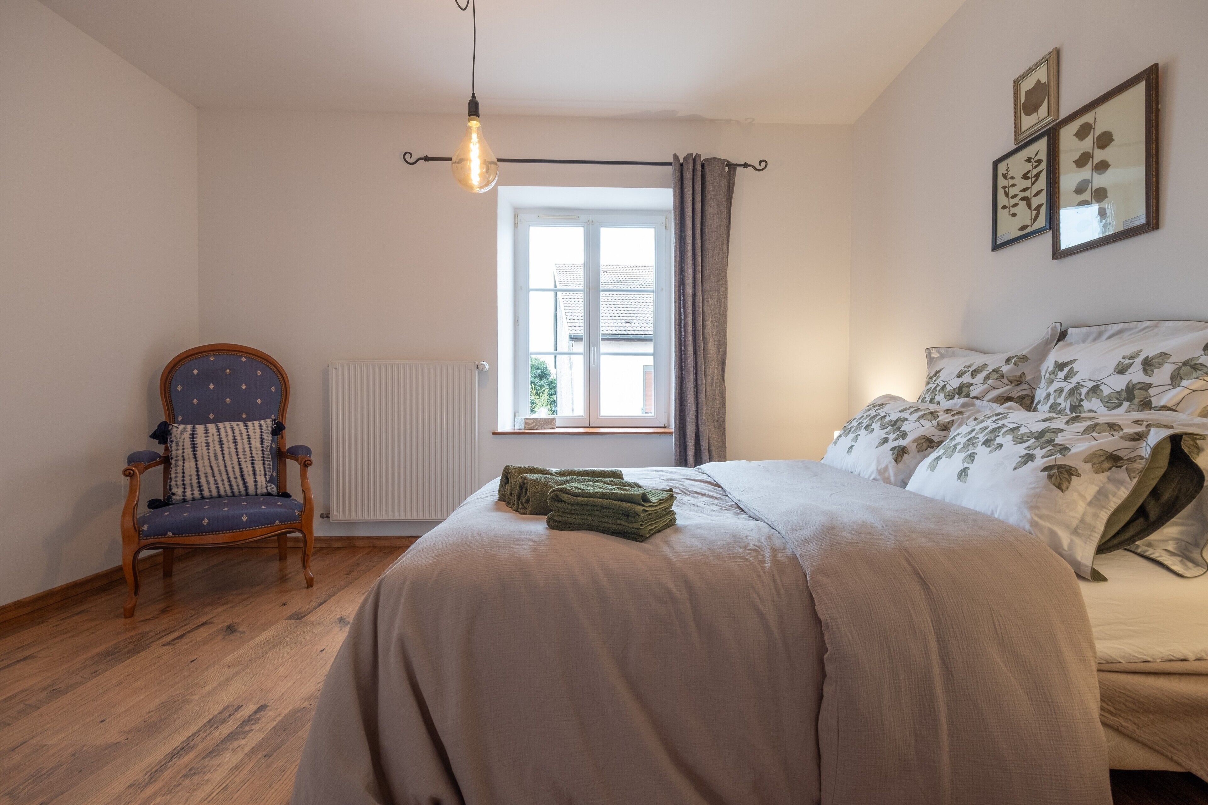 A bedroom with a bed, a chair, a radiator, a window with curtains, and wall art.