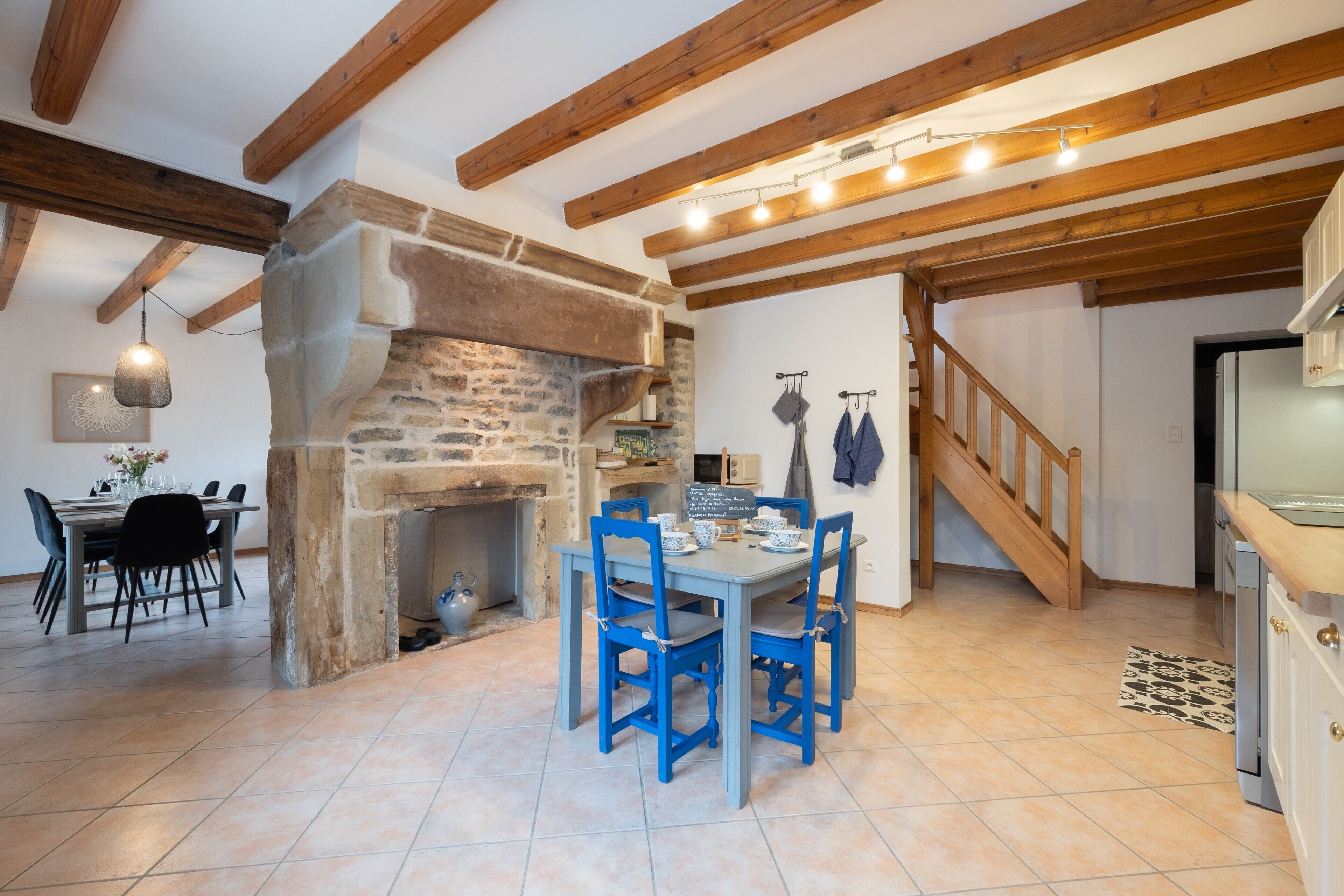 A rustic interior with exposed wooden beams, a stone fireplace, a dining table with blue chairs, and a kitchen area.