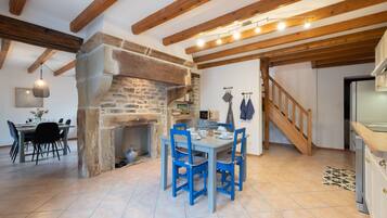 A rustic interior with exposed wooden beams, a stone fireplace, a dining table with blue chairs, and a kitchen area.
