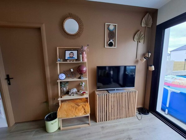 A living room with a TV on a wooden stand, a shelf with various items, a mirror, and a door.