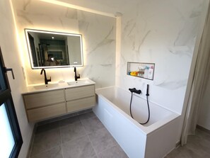 A modern bathroom with a white marble wall, a large mirror, two sinks, a bathtub, and a shelf with toiletries.
