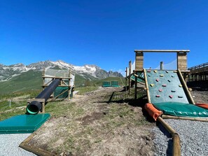 Un parque infantil con estructuras para escalar y un tobogán, con montañas al fondo.