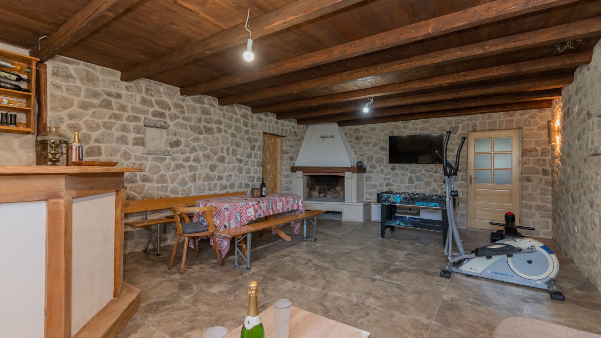 Een rustiek stenen interieur met houten balkenplafond, een stenen openhaard, een tafel met geruit tafelkleed, een bank, een aan de muur bevestigde televisie en een stilstaande hometrainer.