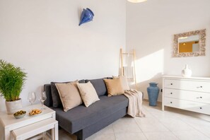 Un salon moderne avec un canapé gris, une table d’appoint blanche ornée d’une plante en pot et de quelques en-cas, un vase bleu et une commode blanche à tiroirs.