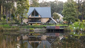A modern house with a large deck, a pond in front, and a forested area in the background. - La Moneta Retreat – A Sanctuary for Healing & Hope -Tranquil Hillside Haven (Dean)