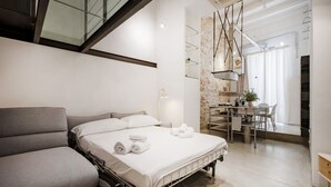 A modern bedroom with a bed, a grey armchair, a dining area with a table and chairs, and a staircase. - Palazzo D’Amelj – Modern Loft in the Heart of Bari Vecchia (Bari)