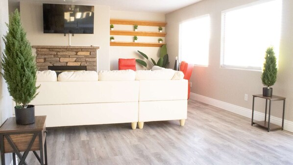 A living room with a white sofa, a stone fireplace, a flat-screen TV mounted above it, and a wooden shelf with small plants.