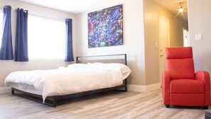 A bedroom with a bed, a red chair, a window with curtains, and a cityscape painting on the wall.