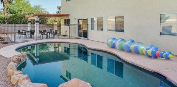 A swimming pool with a curved edge, a covered patio area with outdoor furniture, and a building with multiple windows.