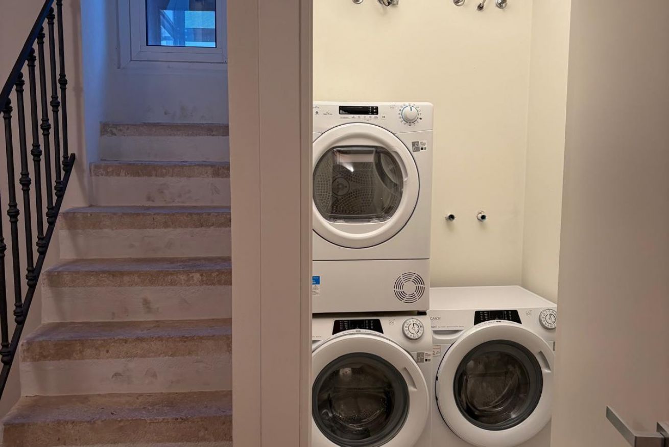A laundry area with two stacked front-loading washing machines and a dryer, a staircase with a black railing, and a small window above the machines.