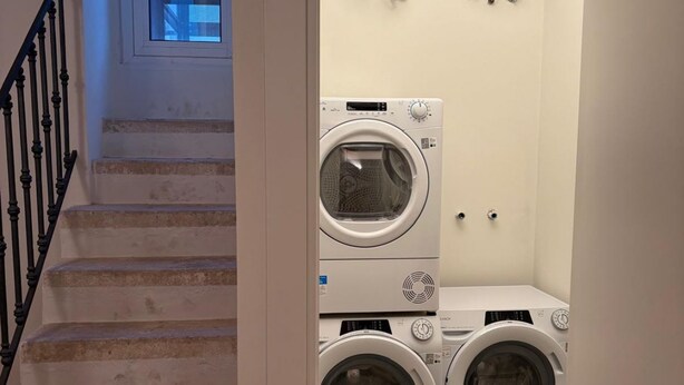 A laundry area with two stacked front-loading washing machines and a dryer, a staircase with a black railing, and a small window above the machines. - Palazzo della Torre 001 - Historical modern stay (Bari)