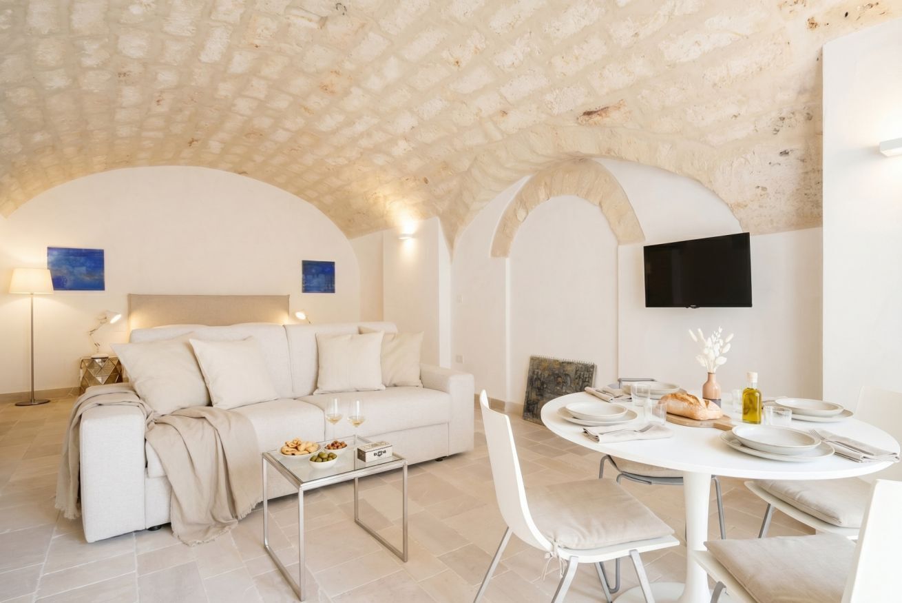 Un soggiorno moderno con un divano bianco, un tavolo da pranzo per due persone e una televisione a schermo piatto fissata al muro.