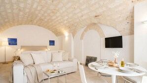 A modern living room with a white sofa, a dining table set for two, and a flat-screen TV mounted on the wall.