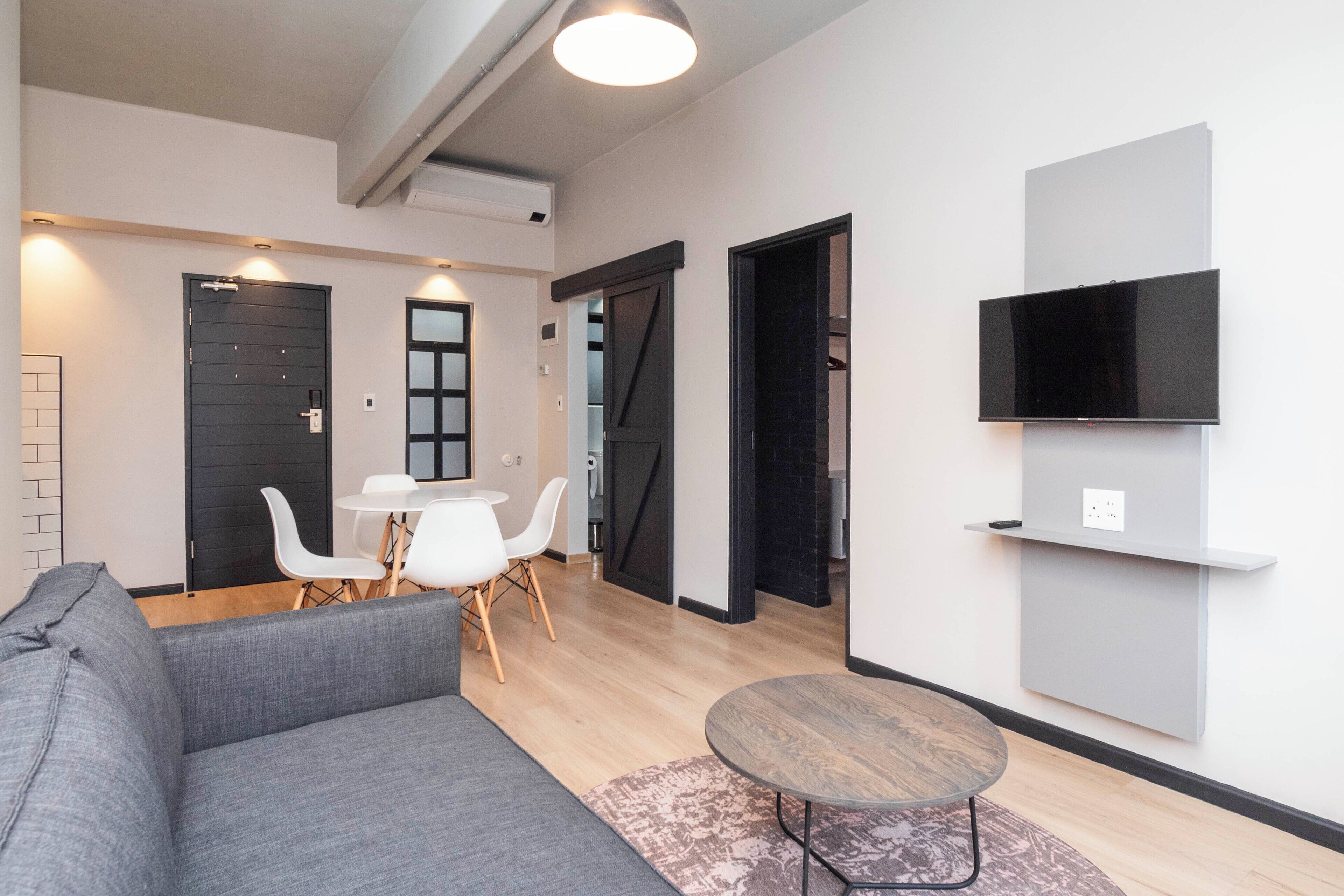 A modern living room with a grey sofa, a round coffee table, and a dining area with a white table and chairs. There is a black door and a black-framed window visible.