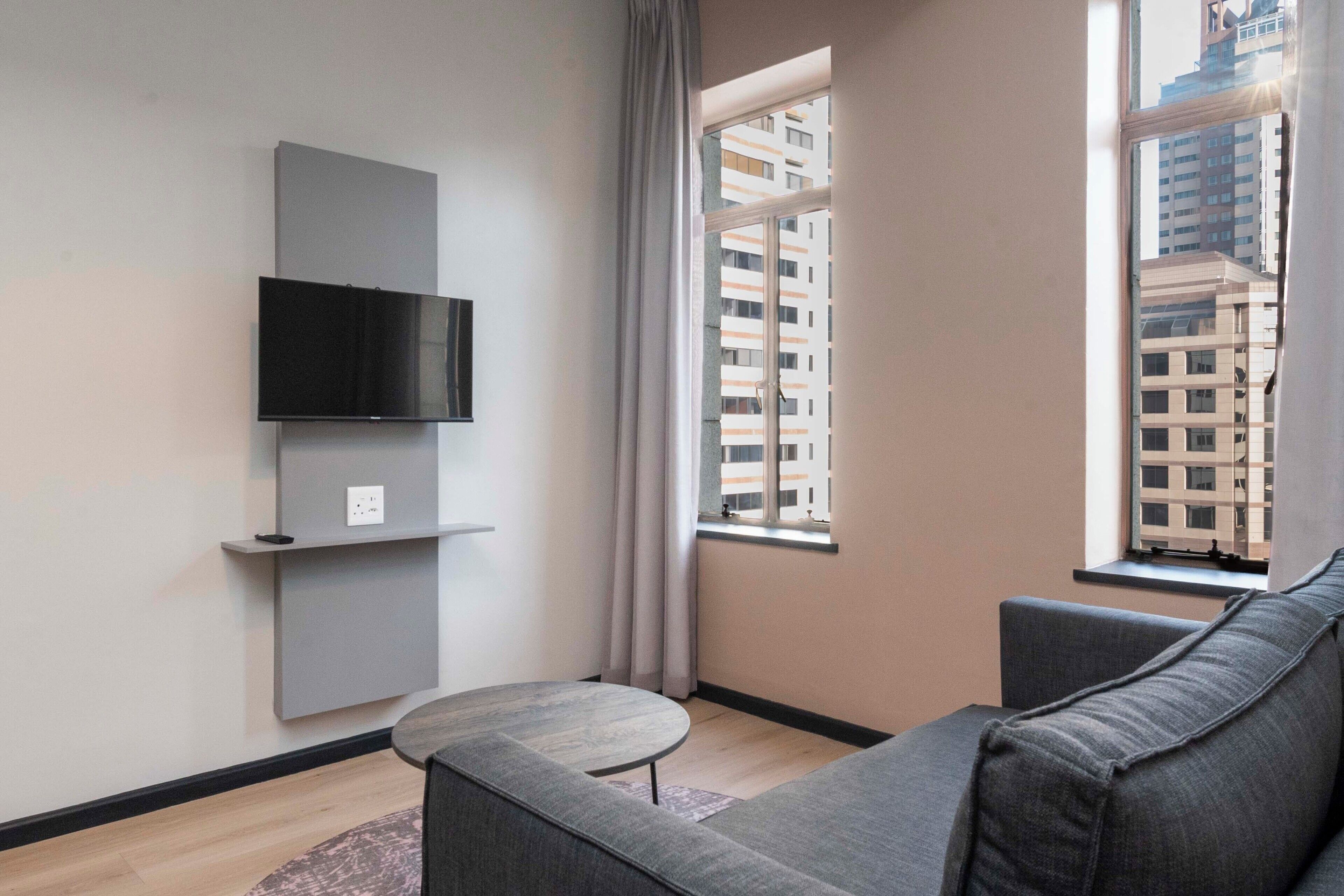 Un salon moderne avec un canapé gris, une petite table ronde, un téléviseur fixé au mur et une fenêtre donnant sur des immeubles.