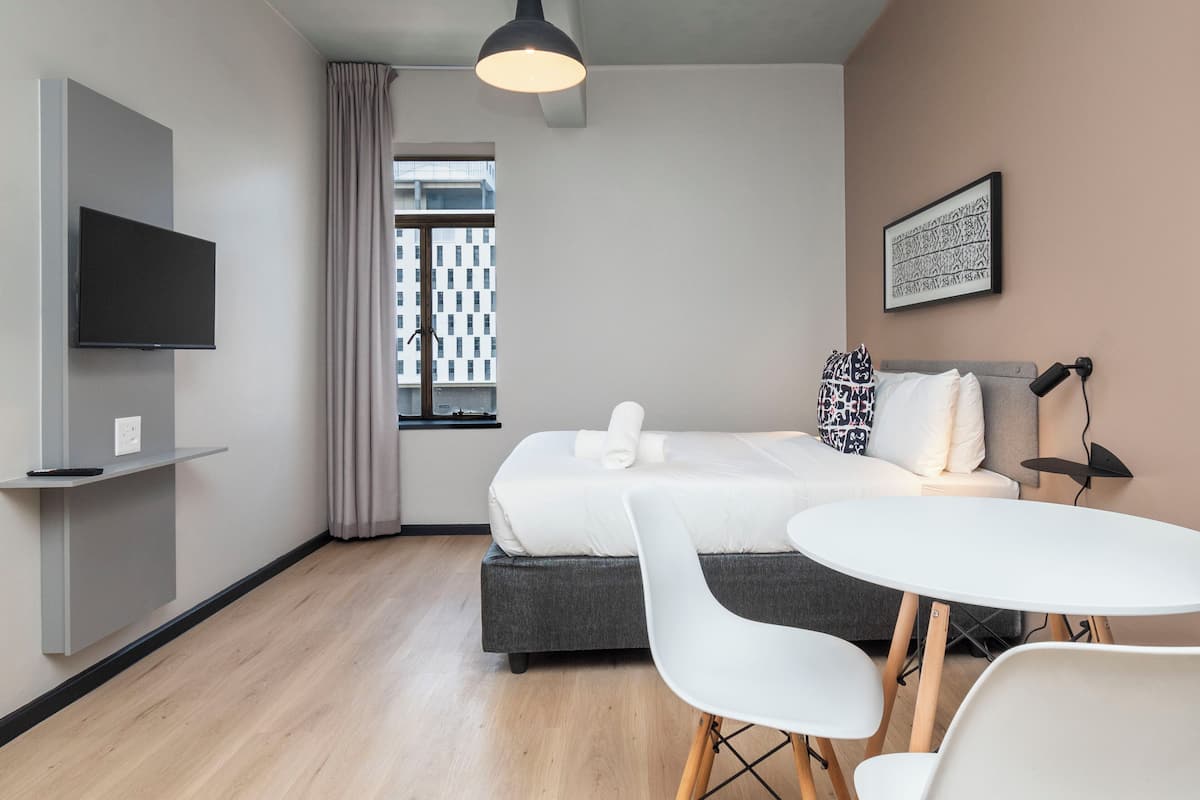 Una habitación de hotel moderna con cama, televisión, mesa de comedor y ventana con cortinas.