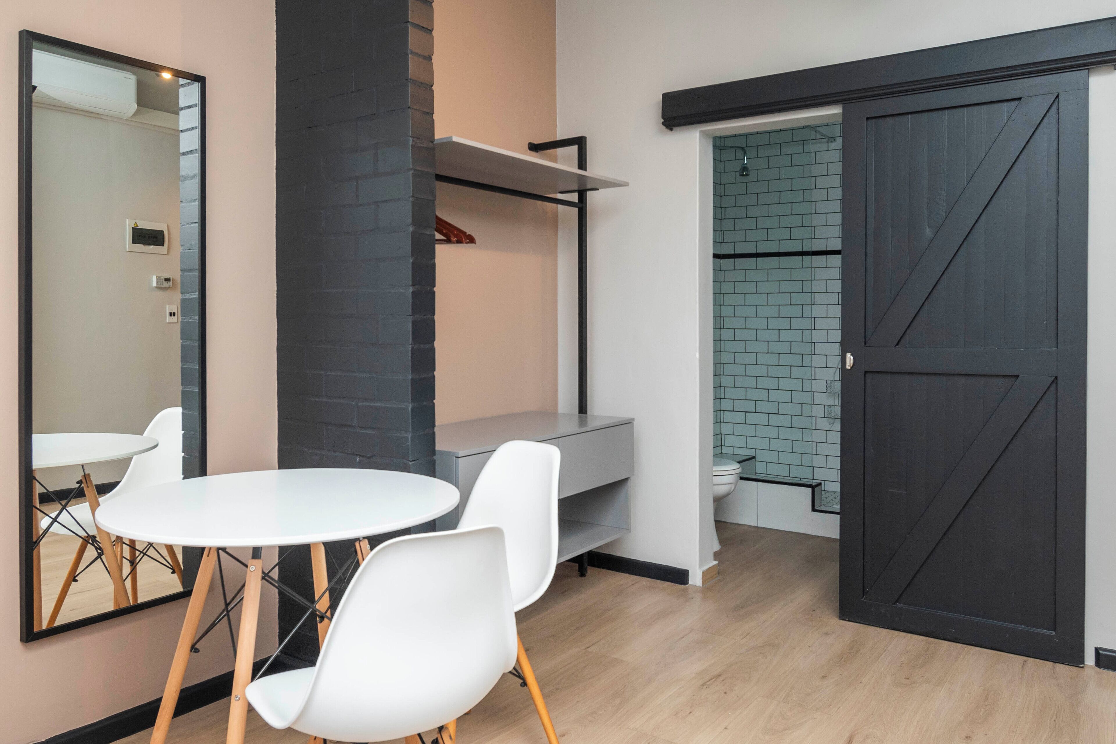 Una habitación moderna con una mesa redonda y sillas blancas, una pared de ladrillo negro decorativa, un banco gris y una puerta corrediza que da a un baño con salpicadero de azulejos.