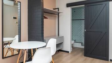 Una habitación moderna con una mesa redonda y sillas blancas, una pared de ladrillo negro decorativa, un banco gris y una puerta corrediza que da a un baño con salpicadero de azulejos.