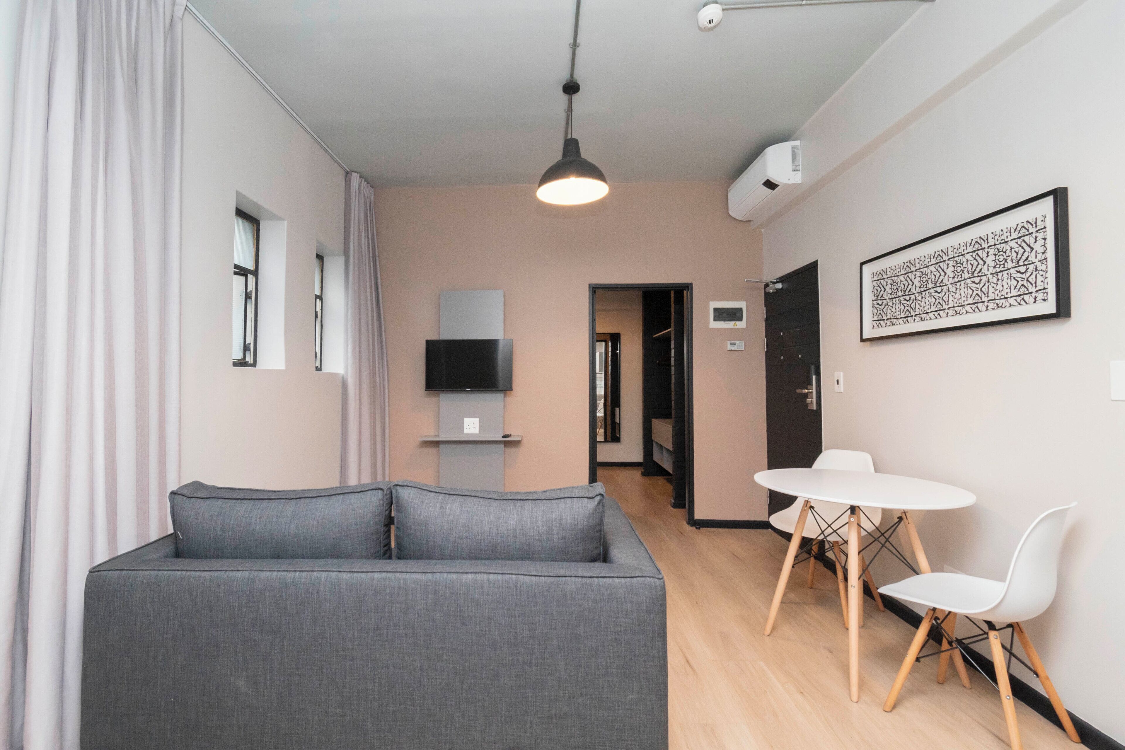 Una sala de estar moderna con un sofá gris, una mesa de comedor blanca con sillas, un televisor de pantalla plana en la pared y una obra enmarcadasobre la mesa.