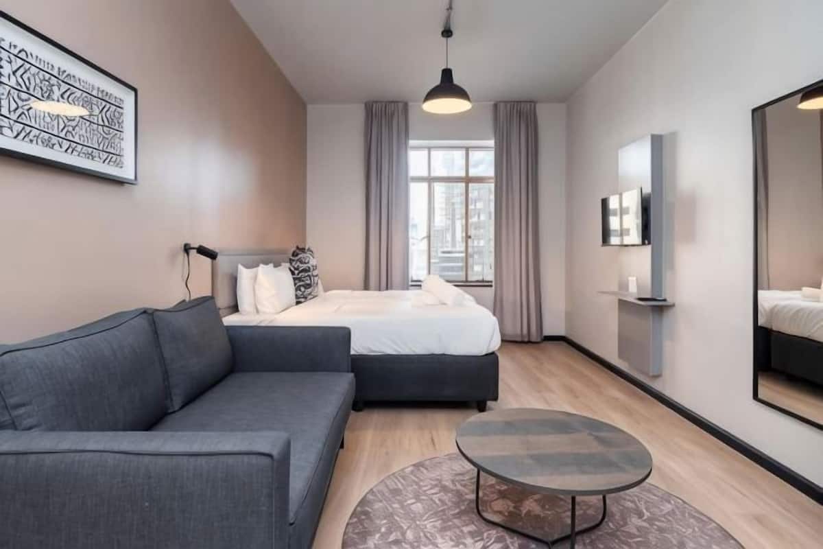 Una habitación de hotel moderna con cama, sofá, una mesita de centro redonda, televisor y un amplio ventanal con cortinas.