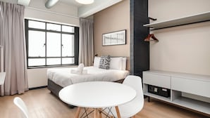 Une chambre d’hôtel moderne dotée d’une grande fenêtre, d’une table de salle à manger ronde, de chaises blanches, d’un lit avec une tête de lit grise, d’une table de chevet blanche et d’une étagère fixée au mur.