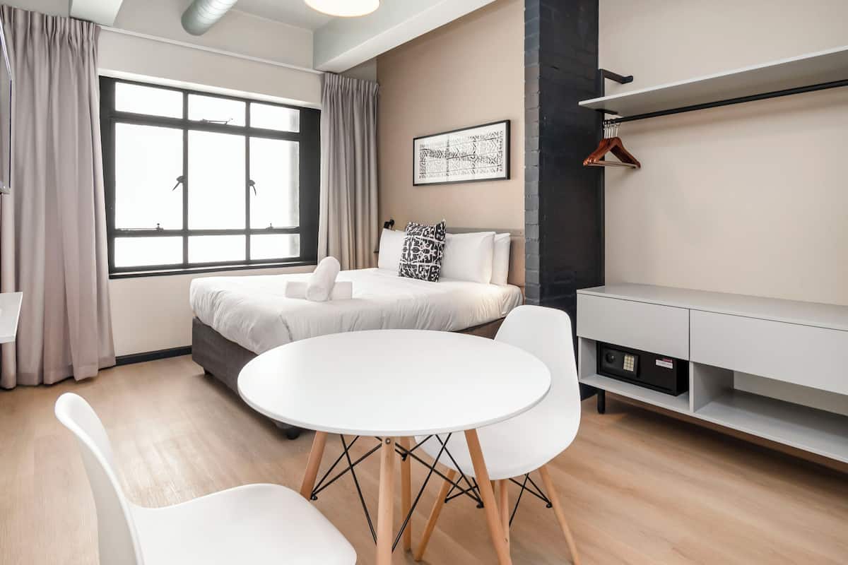 Una habitación de hotel moderna con un ventanal grande, una mesa de comedor redonda, sillas blancas, una cama con cabecero gris, una mesita de noche blanca y un estante fijado a la pared.