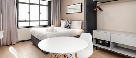 A modern hotel room with a large window, a round dining table, white chairs, a bed with a grey headboard, a white bedside table, and a wall-mounted shelf.