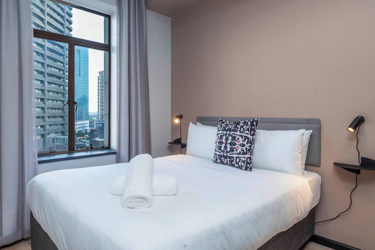Habitación de hotel con una cama grande, una ventana con cortinas, cabecero, mesita de noche con lámpara y vista a la ciudad.