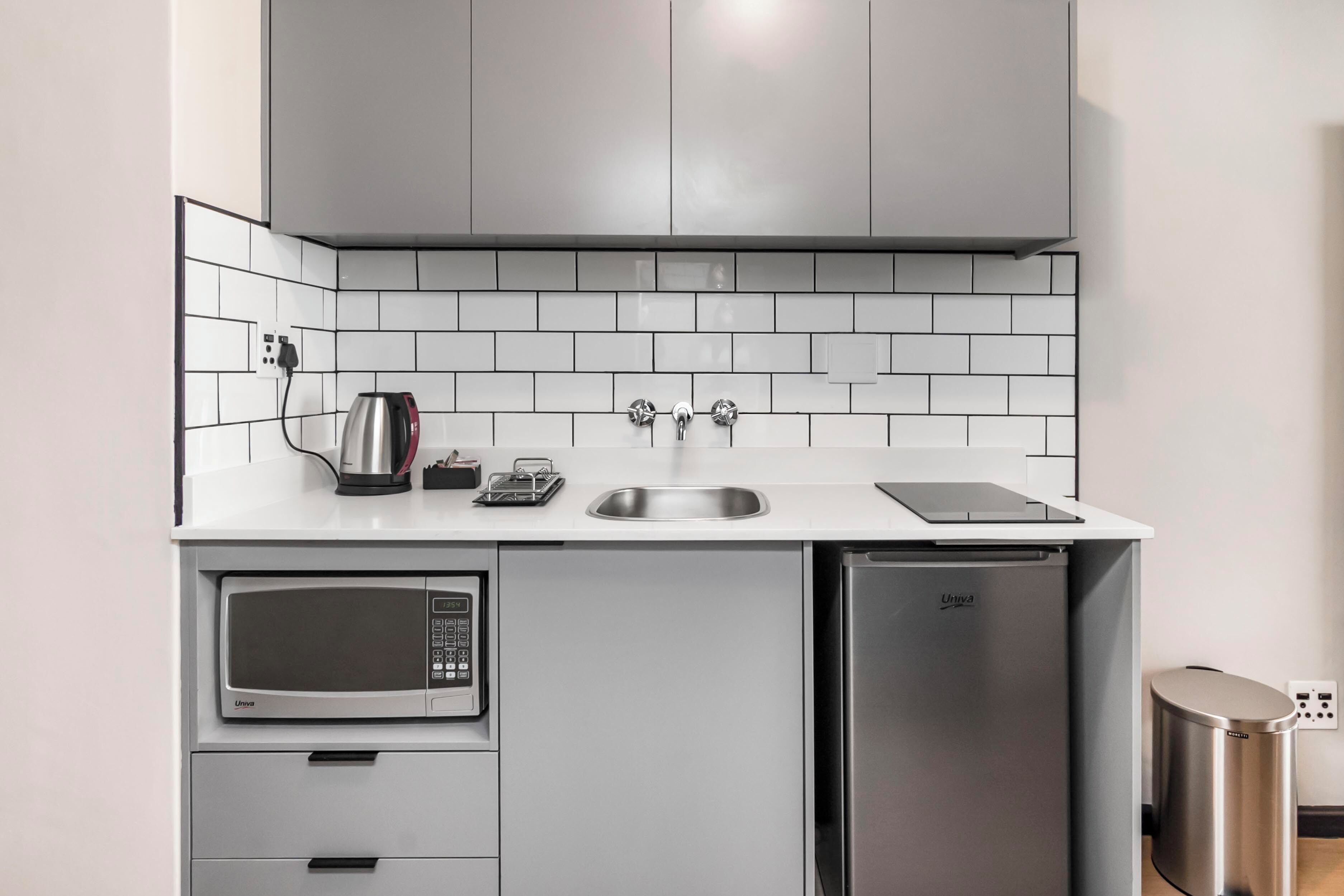 Una cocina compacta con microondas, un refrigerador pequeño, un lavamanos y una tetera.