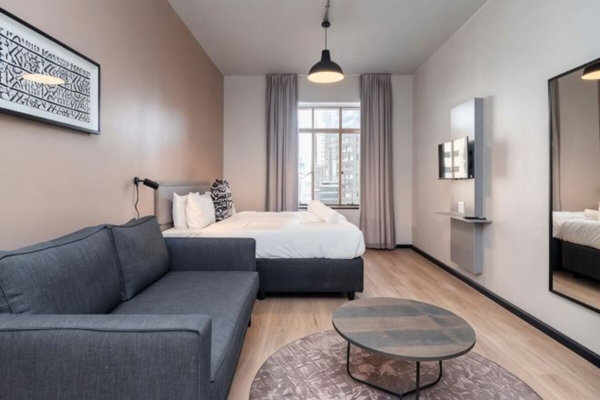 A modern hotel room with a bed, a sofa, a round coffee table, a TV, and a large window with curtains.
