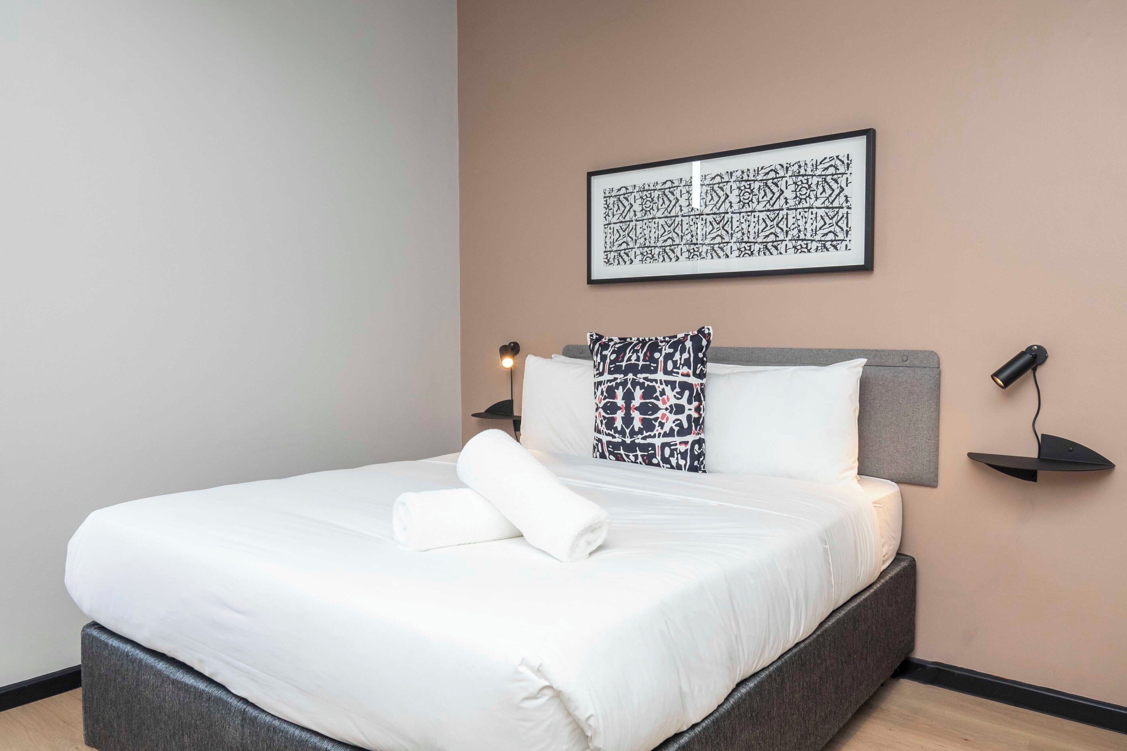 A neatly made bed with white linens and a rolled towel, a patterned pillow, and a wall-mounted abstract artwork.
