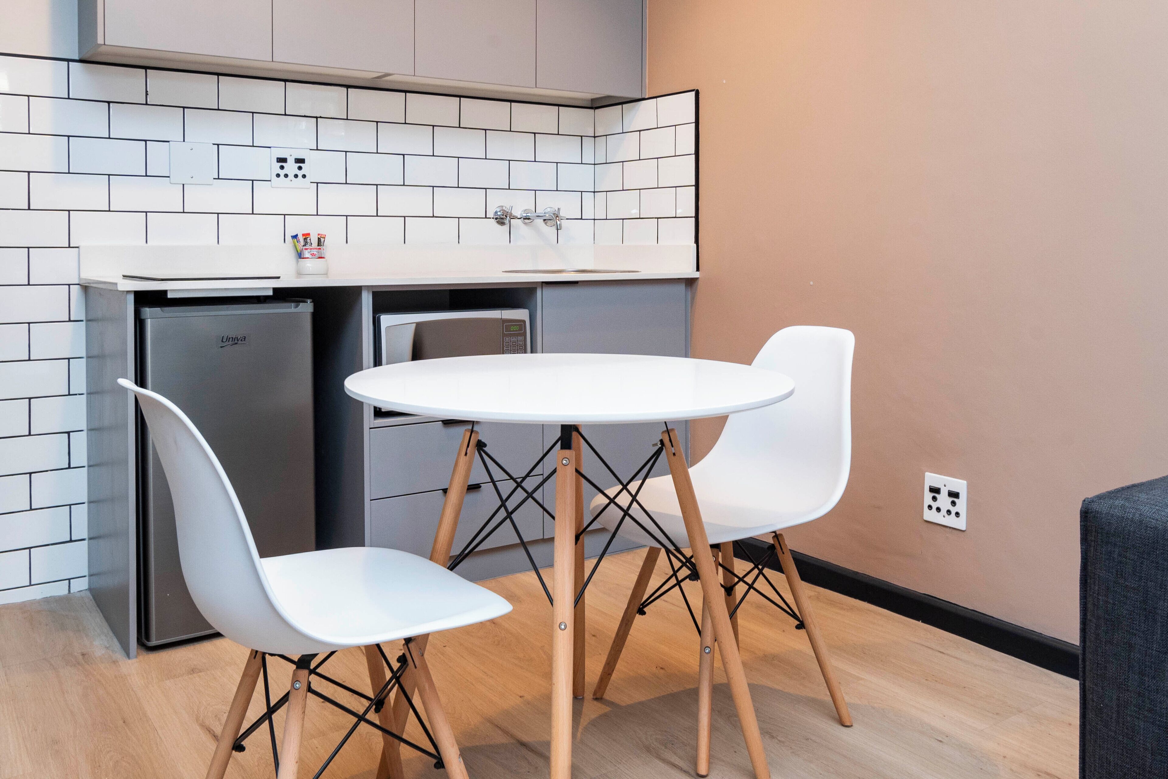 Eine kompakte Küche mit weiß gefliestem Fliesenspiegel, einem eingebauten Kühlschrank, einer Mikrowelle und einem kleinen Spülbecken. Im Hintergrund steht ein runder Tisch mit zwei weißen Stühlen und ein graues Sofa.